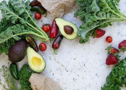 Top view of fresh avocados, kale, tomatoes, and peppers arranged on a marble surface.
